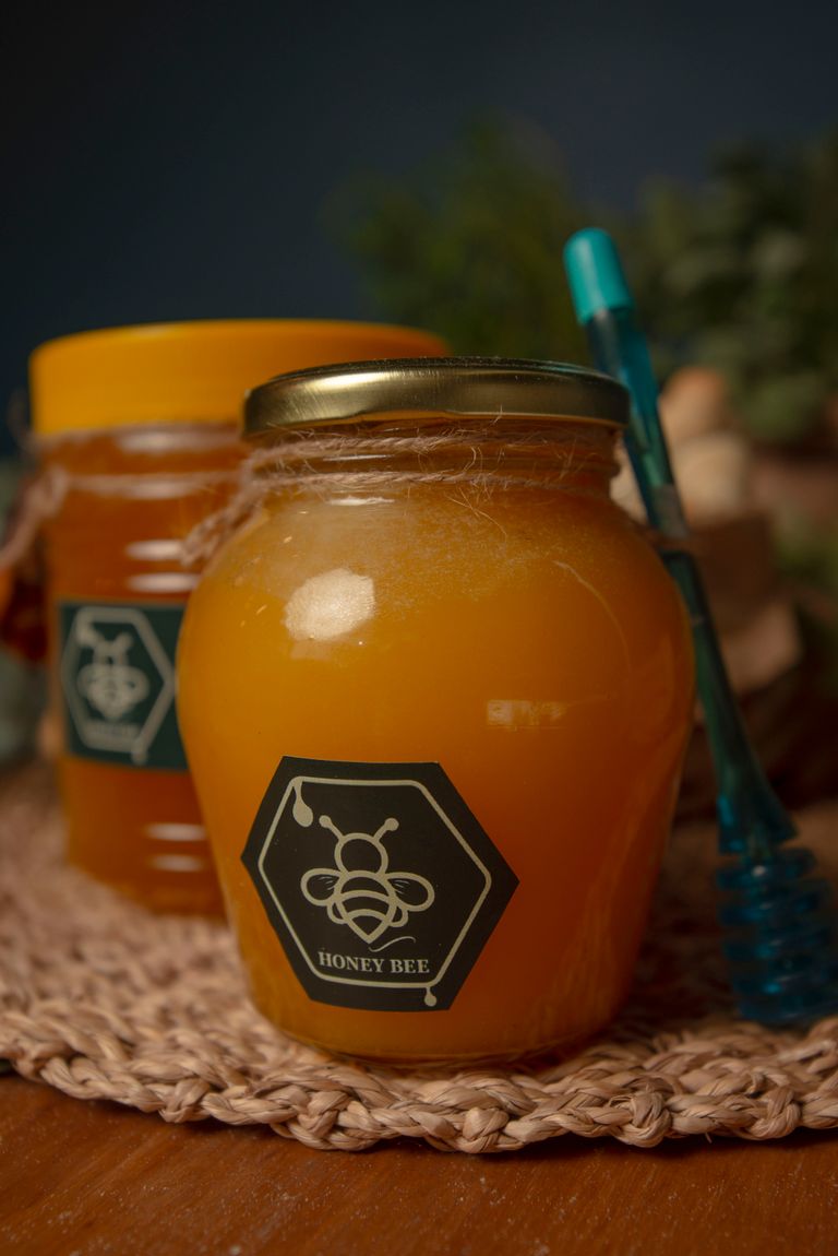 Close-up of a honey jar with hexagonal honeybee label and honey dipper on a wooden surface.