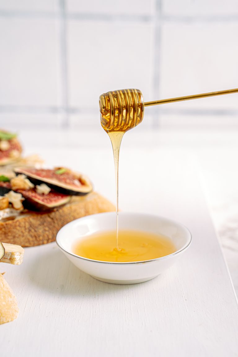 Honey flowing from dipper into bowl beside fig-topped bread on white table.